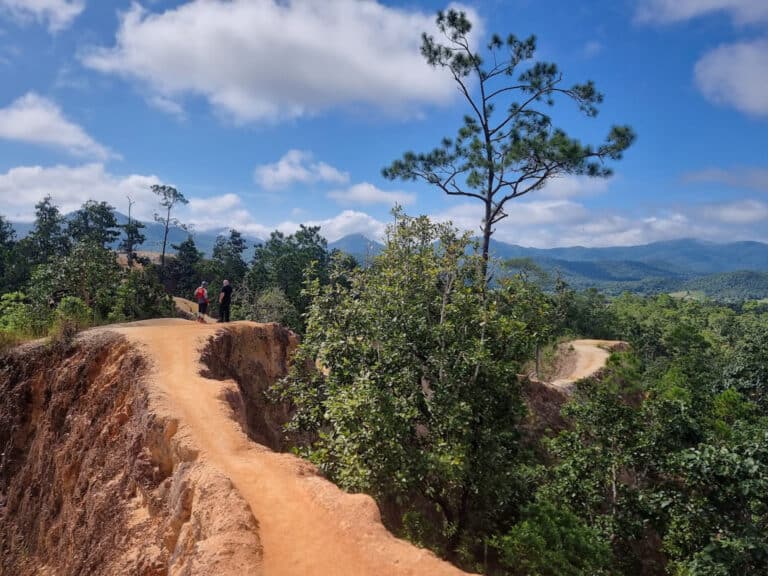 Pai canyon, The image depicts a narrow, winding dirt trail along a steep, eroded cliffside in a lush, forested area. Two people are walking along the path, one wearing a red shirt. The scene is framed by tall pine trees and dense greenery, with a backdrop of rolling hills and mountains under a bright blue sky with scattered clouds. The time appears to be midday, consistent with 01:27 PM +07 on July 02, 2025.