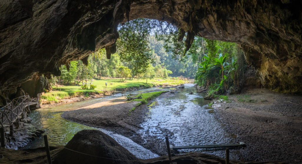 The image depicts Nam Lod Cave, a stunning natural cave located in Thailand. The cave features a beautiful interior with a stream running through it, surrounded by lush greenery visible at the entrance. The scene highlights the cave's rocky walls and the serene environment outside, making it a popular destination for nature enthusiasts.